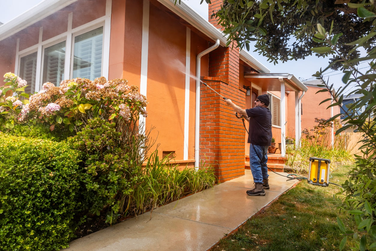 A person uses their DIY skills with a pressure washer to clean the exterior walls of a house with orange siding and brick accents, surrounded by plants and shrubs on a sunny day.