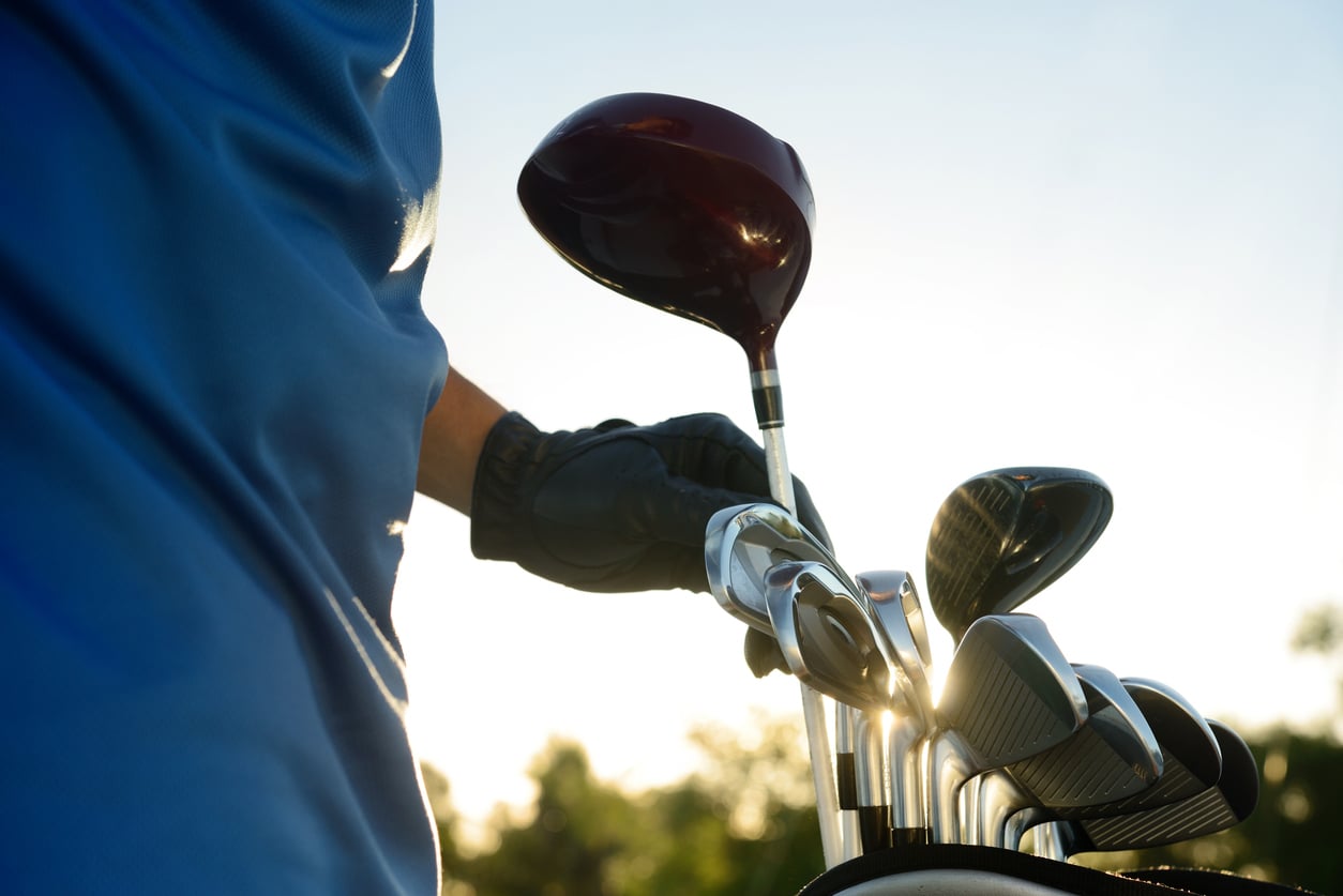 A person wearing a blue shirt and black glove is selecting a golf club from a golf bag, highlighting products men skip buying, with sunlight shining in the background.