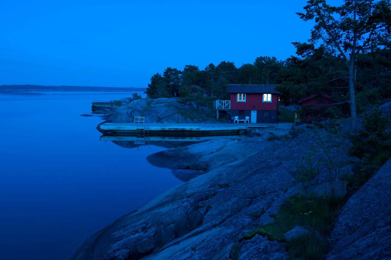A small red cabin with lit windows sits on rocky shoreline beside a calm blue lake at dusk, surrounded by trees and a simple wooden dock.