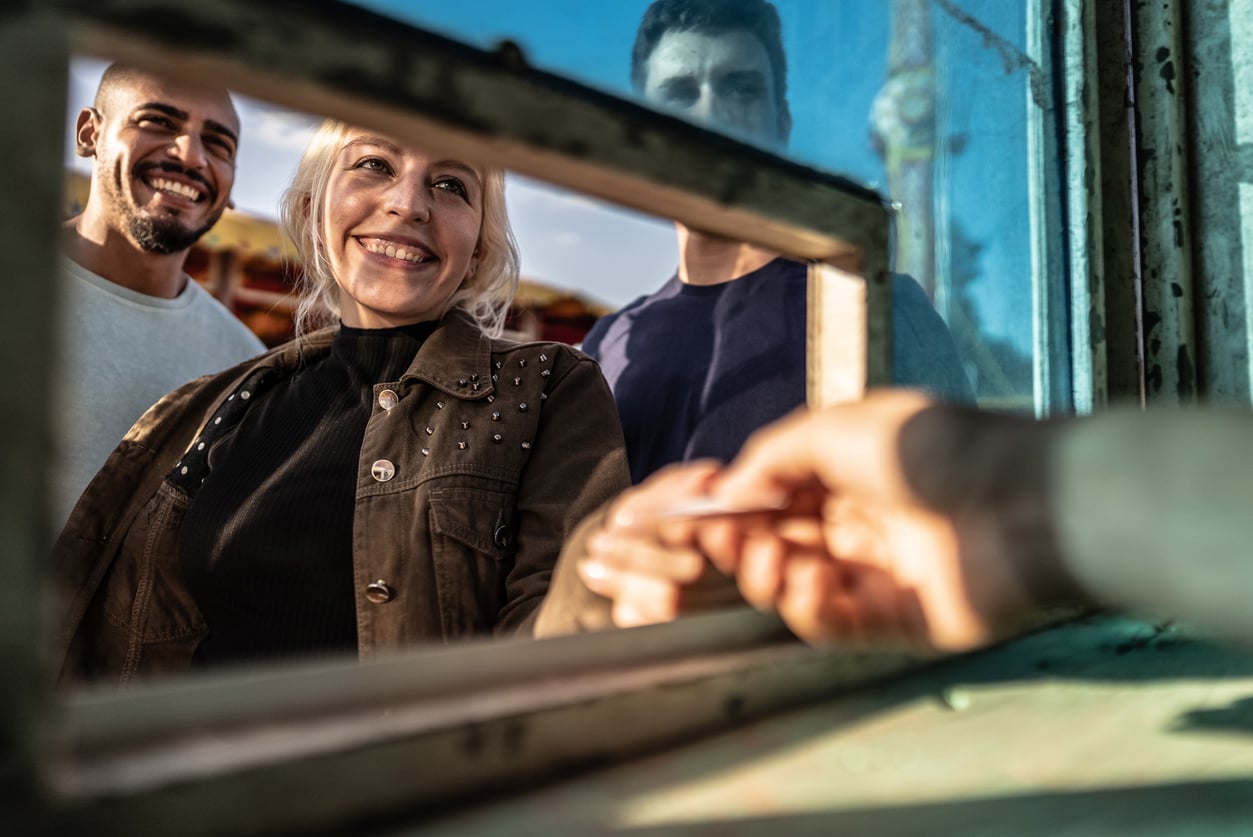 Three people stand outside a ticket booth window, smiling as one of them uses clever concert ticket hacks to hand over a ticket or money through the window. The scene is bright and cheerful.