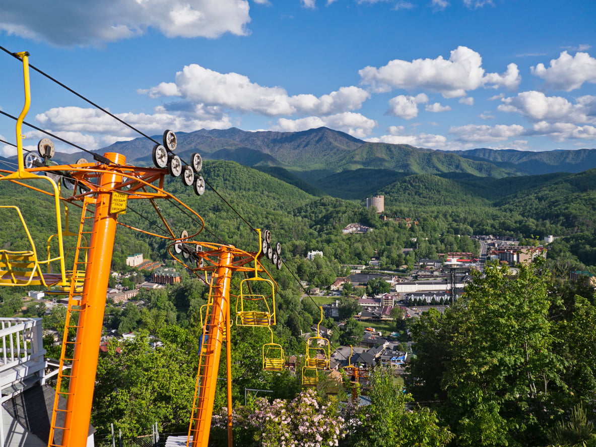 Bright orange chairlift rises over a green, forested landscape with a small town below and mountains in the background under a blue sky dotted with white clouds—an inviting escape for those seeking Florida alternatives.