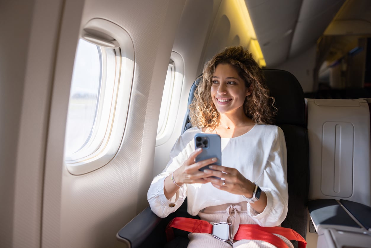 A woman with curly hair sits by the window on an airplane, smiling and holding a smartphone. She is wearing a white top and red seatbelt, and sunlight shines through the window onto her.