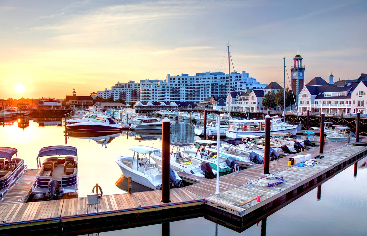 A marina at sunset with several boats docked along wooden piers, calm water reflecting the sky, and modern waterfront buildings and a lighthouse-style structure in the background.