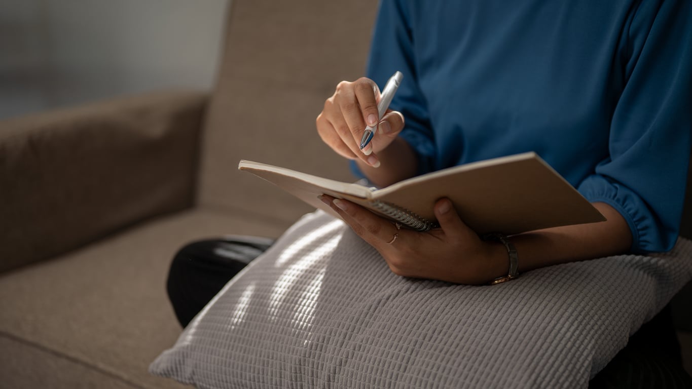 A person sitting on a couch with a cushion, holding a notebook and pen, appears to be writing or taking notes—perhaps journaling as part of their mental health routine. The focus is on their hands and the notebook.