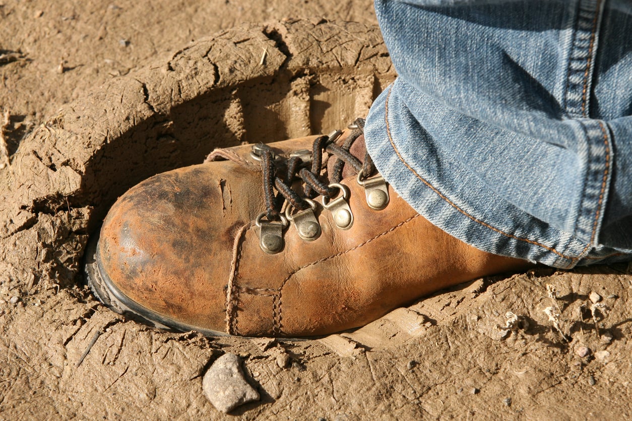 A close-up of a worn brown hiking boot—one of the classic products men still buy—stepping into a large footprint in dry, cracked dirt, with the person wearing blue jeans.
