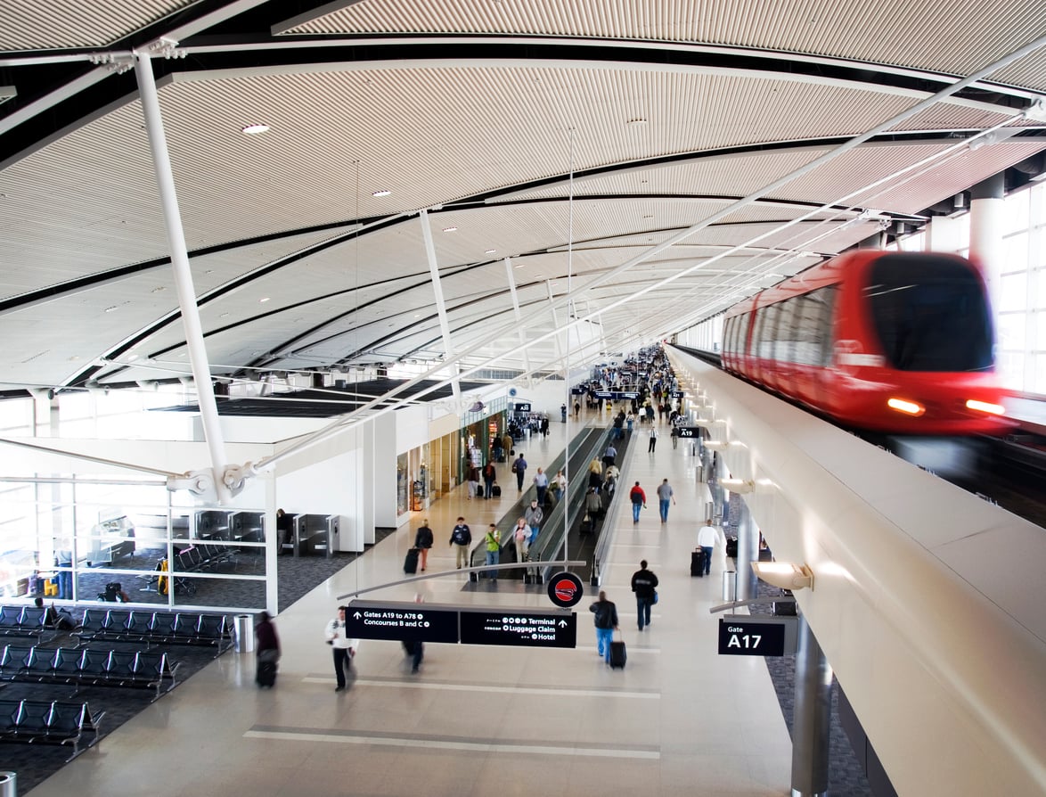 Wide view of a modern, convenient airport terminal with travelers walking, seated areas, shops, and a red automated train moving on an elevated track above the main concourse. Signs indicate directions to different gates.