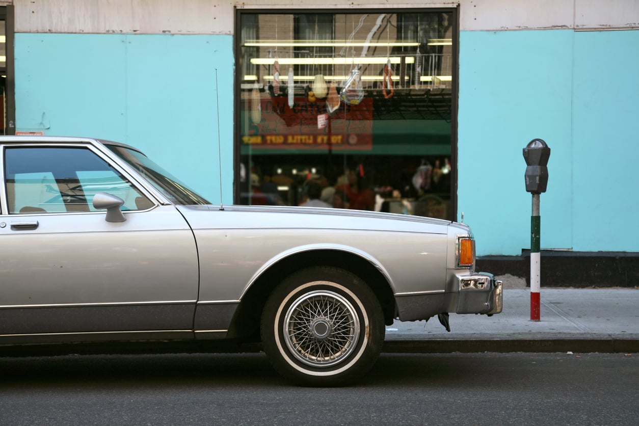 A silver vintage car is parked on the street beside a parking meter with the colors of the Mexican flag. Behind the car is a building with turquoise walls and a window displaying items inside.