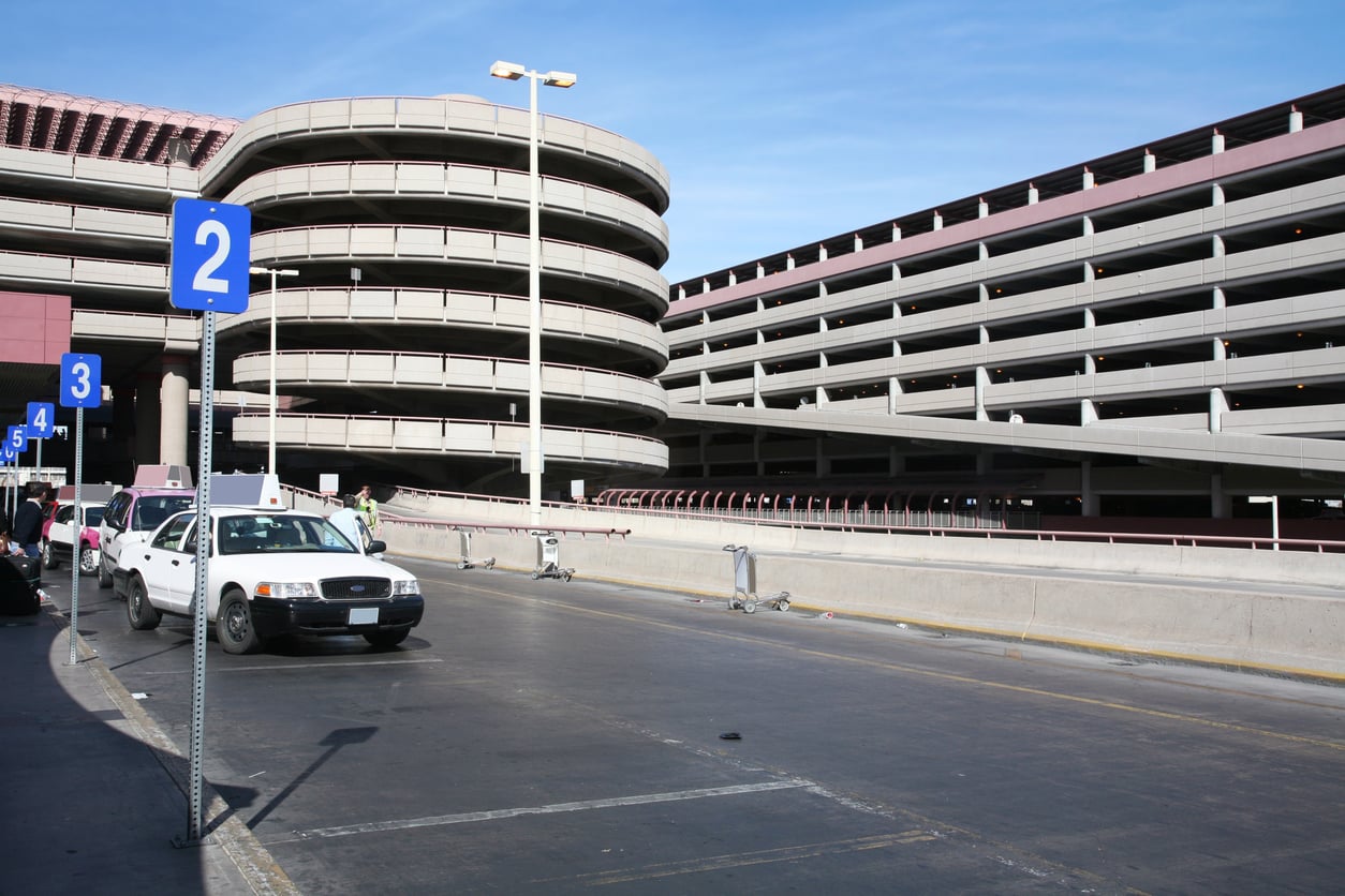 A multi-level parking garage with circular ramps is shown—ideal for visitors seeking Vegas money-saving tips. Several white taxis wait by the curb under blue number 2 signs, while a few people and shopping carts gather on the sidewalk.