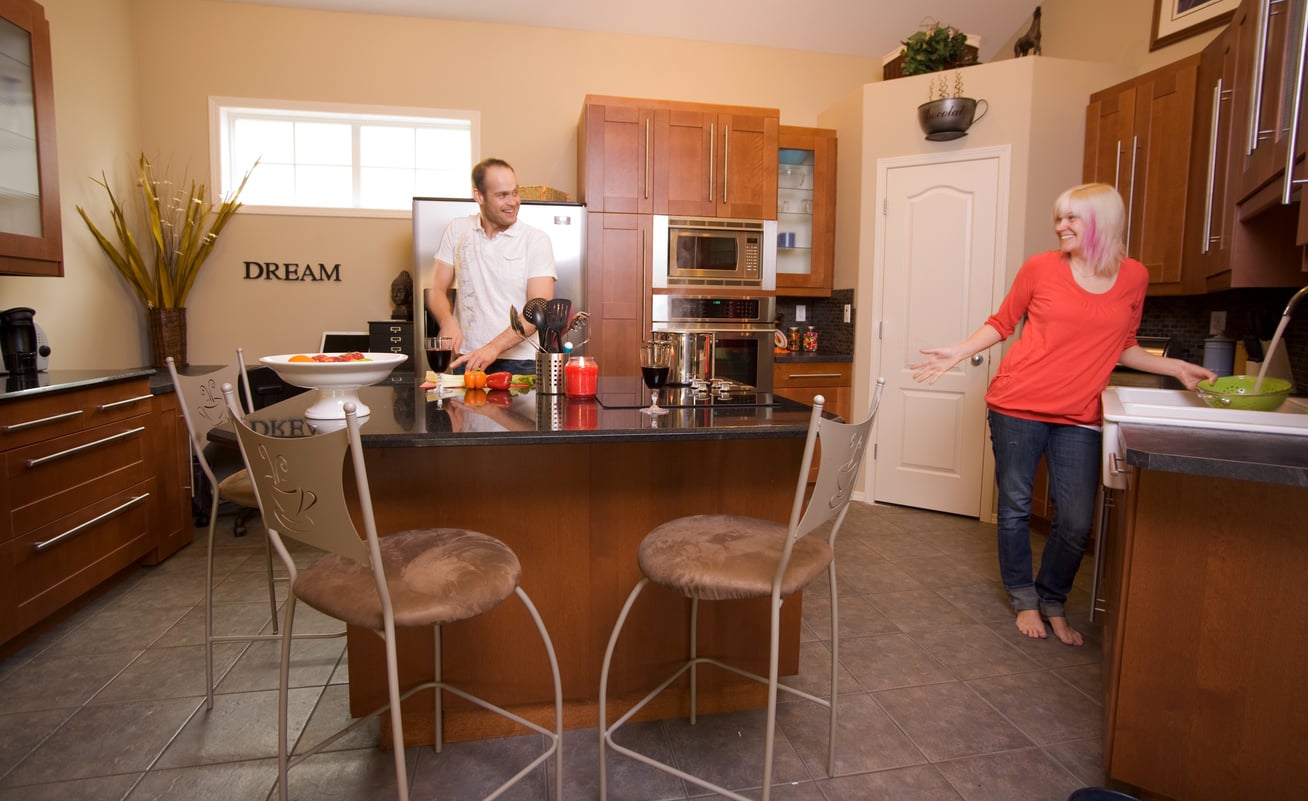 A man stands by a kitchen island preparing food while a woman in a red shirt smiles and gestures from near the sink in a modern kitchen with wooden cabinets.