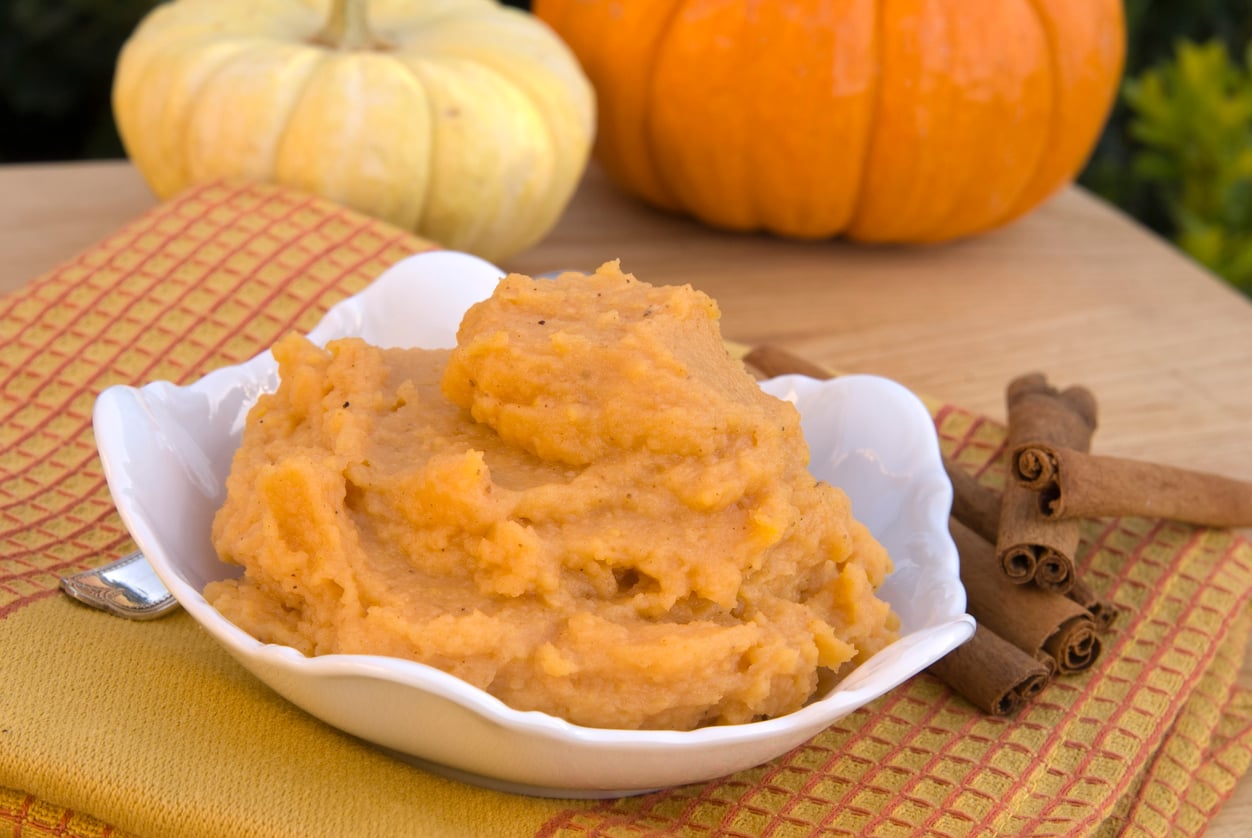 A bowl of mashed sweet potatoes sits on a yellow and orange checkered cloth, with cinnamon sticks beside it, two pumpkins in the background, and perfect for recipes needing egg substitutes.
