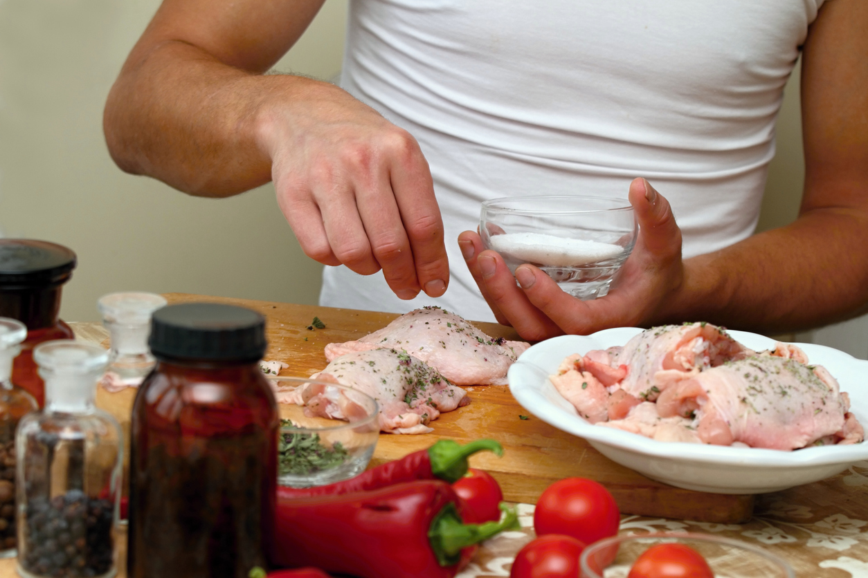 A person sprinkles salt onto raw chicken pieces on a cutting board, surrounded by spices, herbs, tomatoes, and red peppers in a kitchen setting.