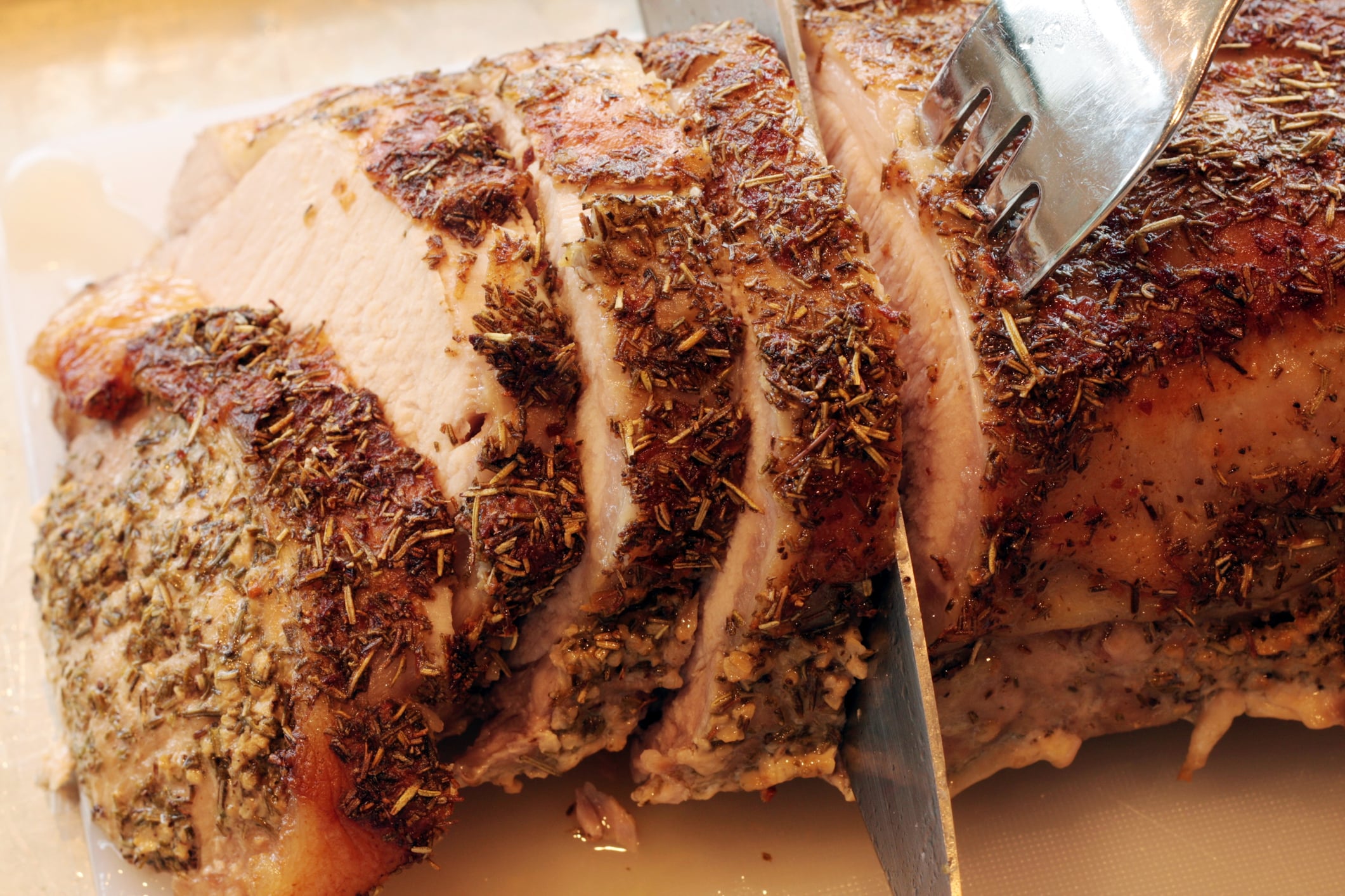 Cheap foods that are expensive now. Close-up of a roasted, herb-crusted pork loin being sliced with a knife and held in place by a fork. The meat is juicy and tender, showing a golden-brown crust seasoned with herbs.