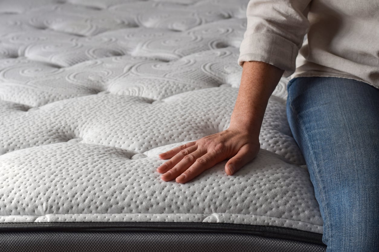 A person wearing jeans and a light shirt sits on the edge of a white, textured mattress, pressing one hand gently into its surface to test its softness—perfect for those considering delayed purchases and wanting assurance before deciding.