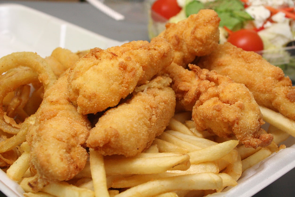 A takeout container filled with golden fried chicken tenders, crispy French fries, and onion rings, with a fresh salad topped with tomatoes and feta cheese in the background.