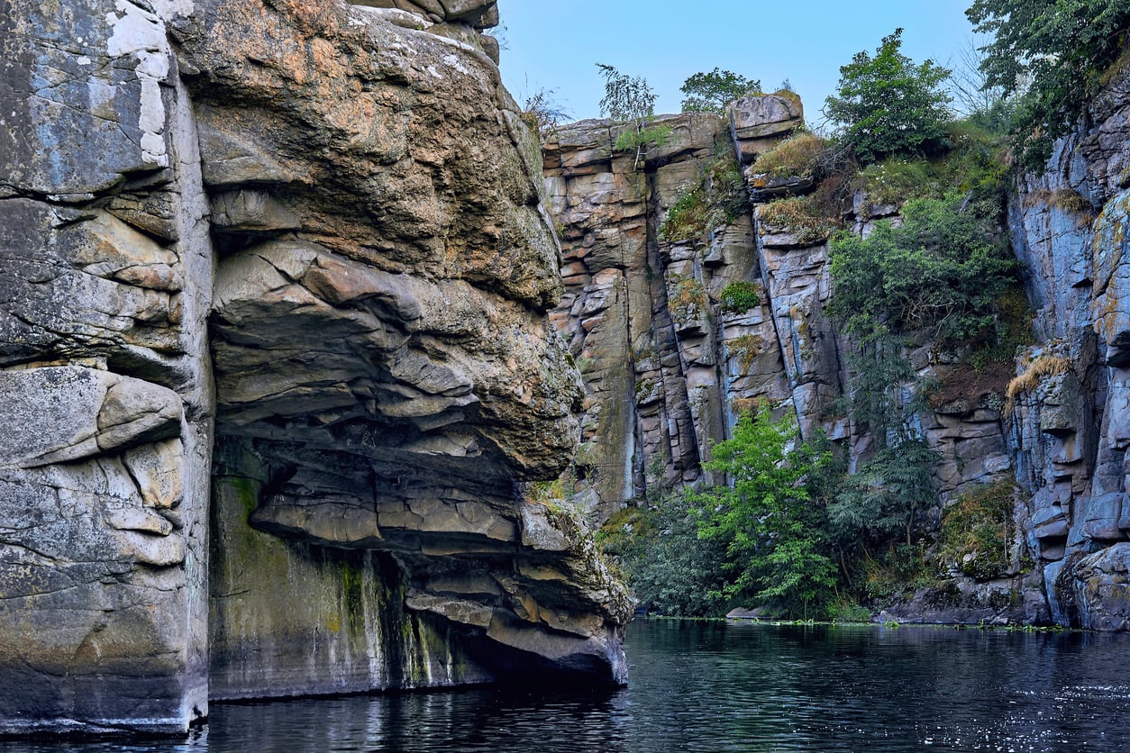 Tall, rugged rock cliffs rise above a calm river, with patches of green vegetation and trees growing along the rocky surfaces and at the water's edge under a clear blue sky.