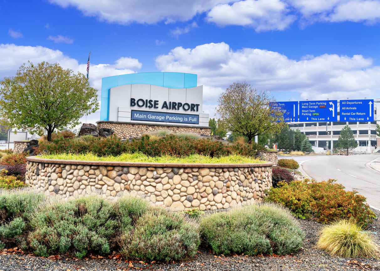 Entrance sign for Boise Airport, one of the most convenient airports, surrounded by landscaped greenery and a stone wall, with trees and directional signs for parking and departures in the background under a partly cloudy sky.