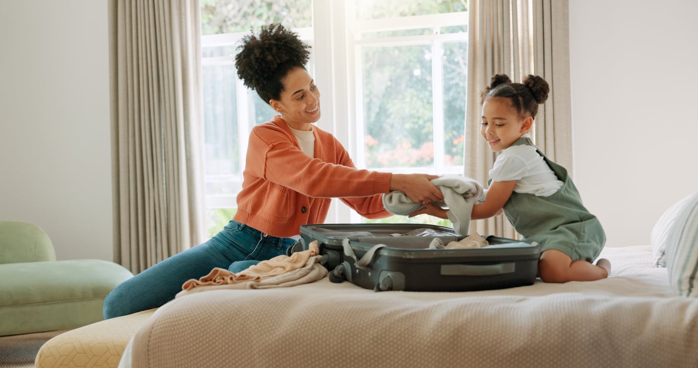 A woman and a young girl sit on a bed, smiling as they pack clothes into an open suitcase together. Sunlight streams through large windows behind them, creating a warm, cheerful atmosphere that reflects their focus on hotel room safety.
