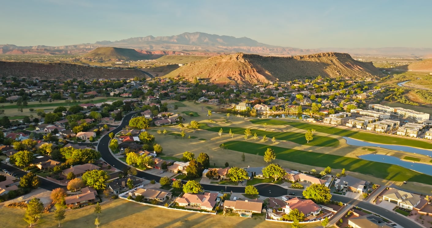 Aerial view of a suburban neighborhood with scattered houses, green lawns, golf course, and winding roads, set against rocky hills and distant mountains under a clear sky.