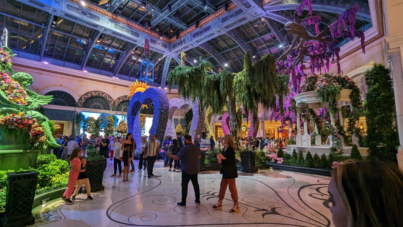 People explore a large indoor garden with elaborate floral sculptures, greenery, and colorful lights under a glass ceiling—a vibrant Las Vegas attraction that’s perfect for visitors seeking budget tips while enjoying stunning displays and snapping photos.