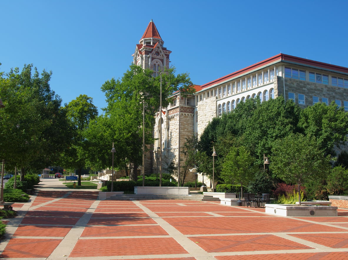 A wide brick plaza with trees and benches leads to a historic stone building with a clock tower under a clear blue sky, capturing the inviting charm found in many affordable college towns.
