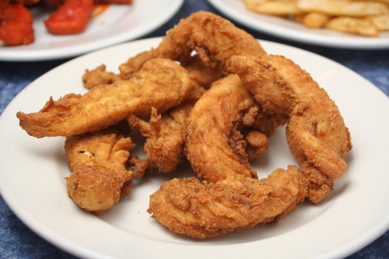 A white plate filled with several pieces of golden-brown fried chicken tenders, set on a table with other plates of food visible in the background.