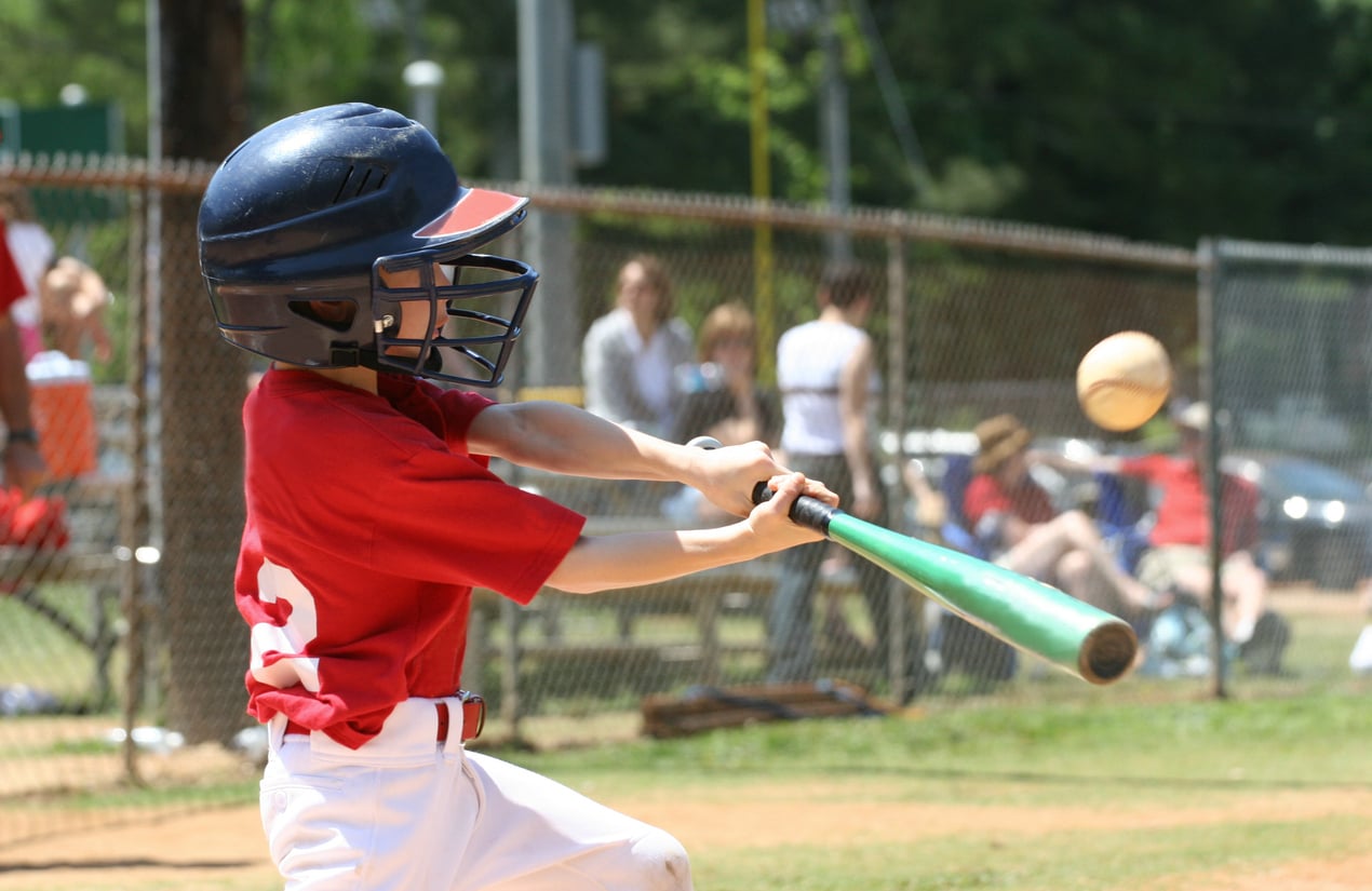 A young baseball player in a red jersey and helmet swings a green bat at an incoming baseball on an outdoor field, with spectators sitting on benches behind a chain-link fence. Cost of living in 1995.