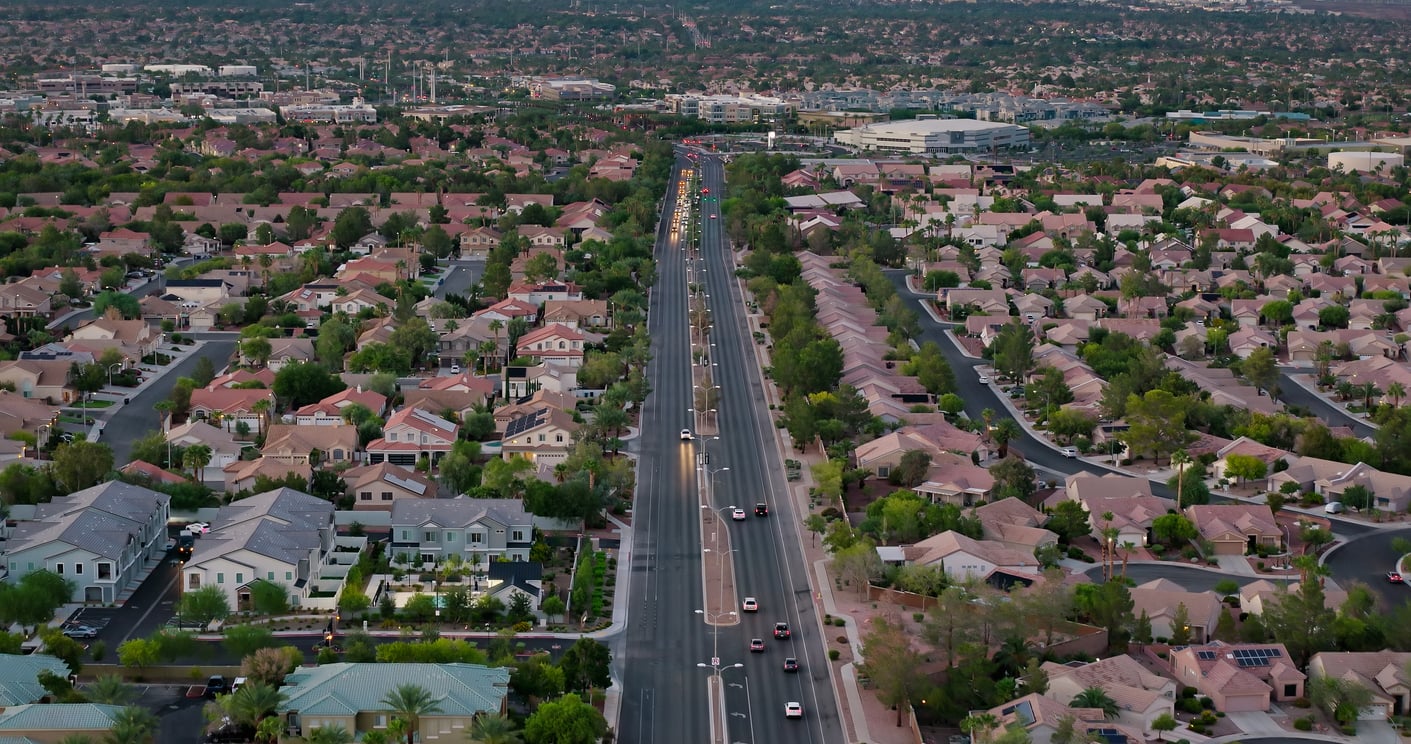 A wide aerial view of a suburban neighborhood with rows of houses on both sides of a busy main road, cars driving in both directions, and green trees scattered throughout the area.