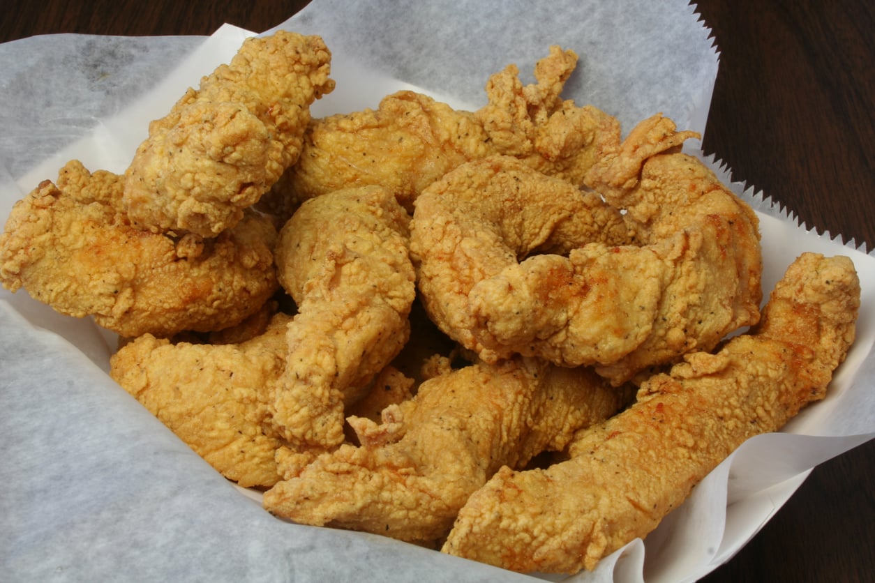 A basket lined with white parchment paper filled with several pieces of golden-brown fried chicken tenders on a dark wooden surface.