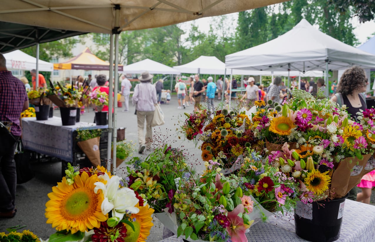 Colorful flower bouquets displayed on tables at an outdoor market, showcasing vibrant small business states, with people walking among white vendor tents in the background on a bright, clear day.