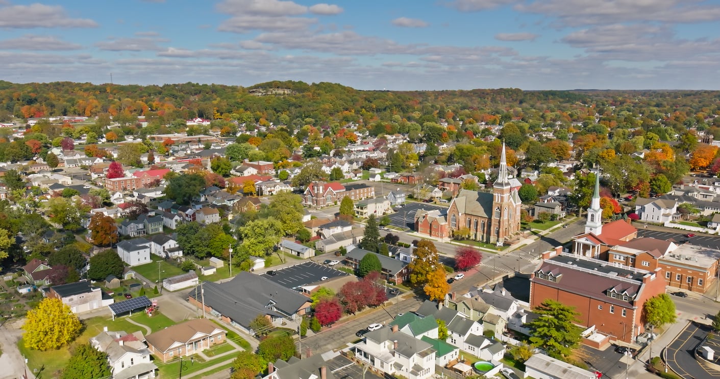 Aerial view of a small town with tree-lined streets, houses, churches with tall steeples, and autumn foliage under a partly cloudy sky.