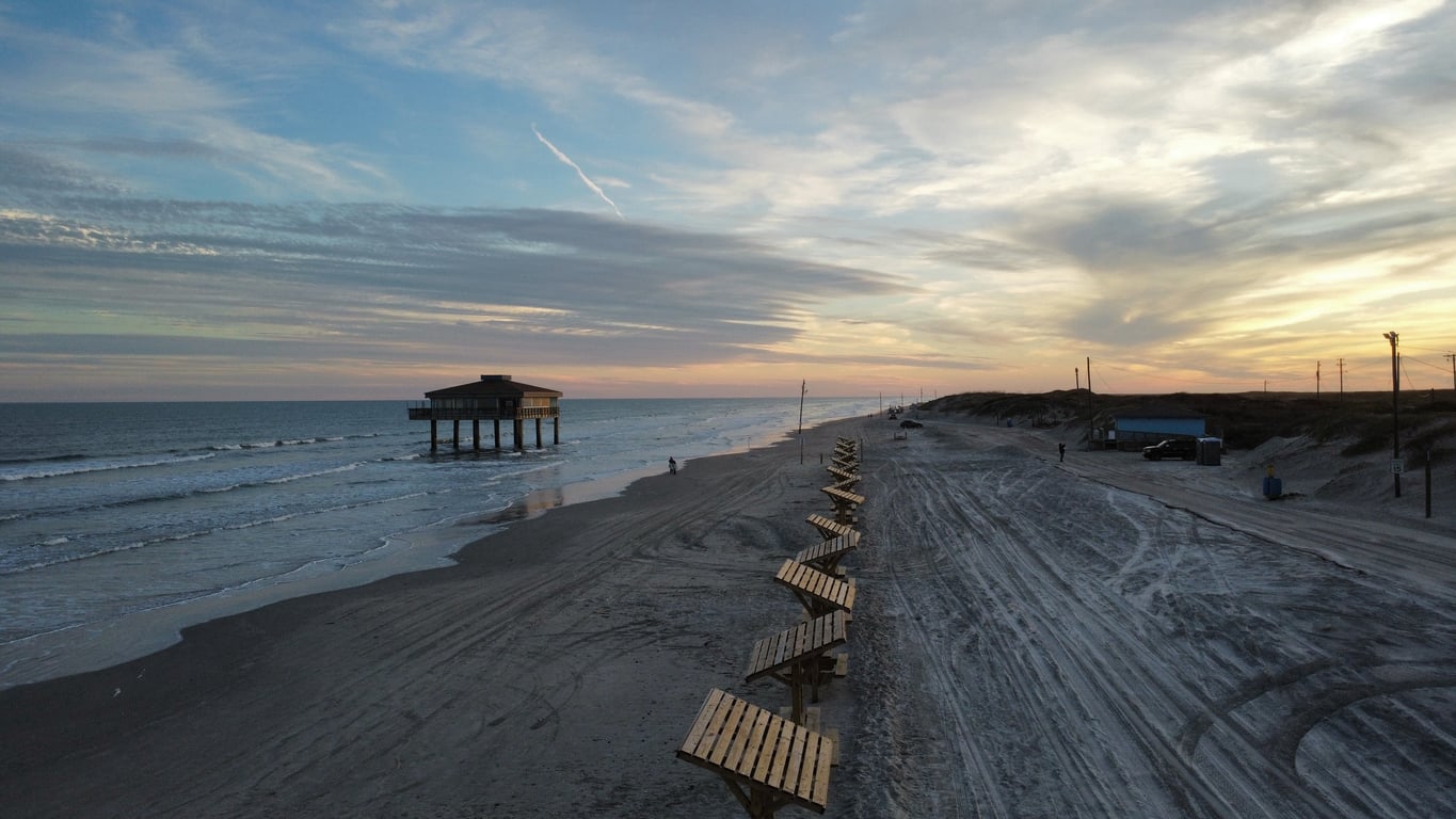 A quiet beach at sunset with gentle waves, a pier house on stilts over the water, and a row of empty benches along the sandy shore. The sky is streaked with clouds, and the scene feels calm and peaceful.