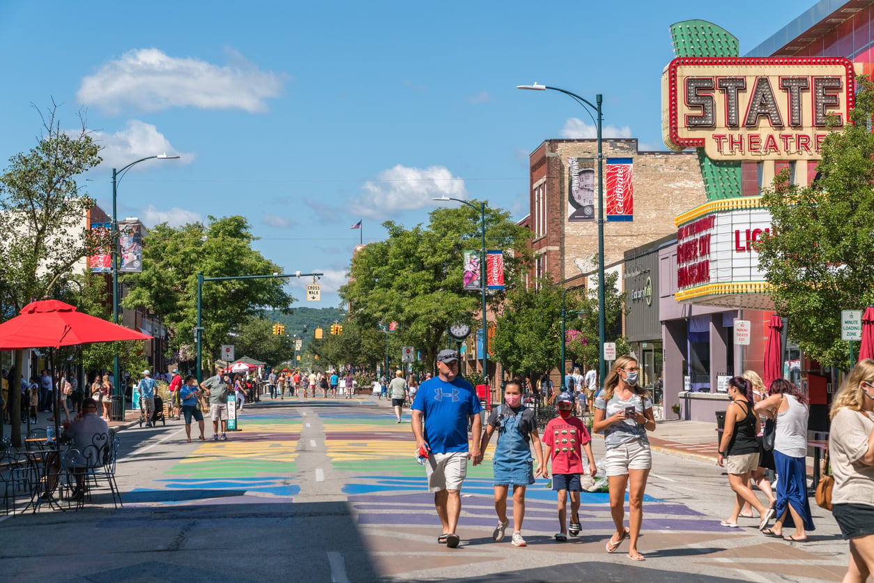 People walk on a colorful, pedestrian-friendly street lined with shops, cafes, and trees on a sunny day. A large marquee reads "State Theatre" on the right side. The atmosphere is lively and relaxed.