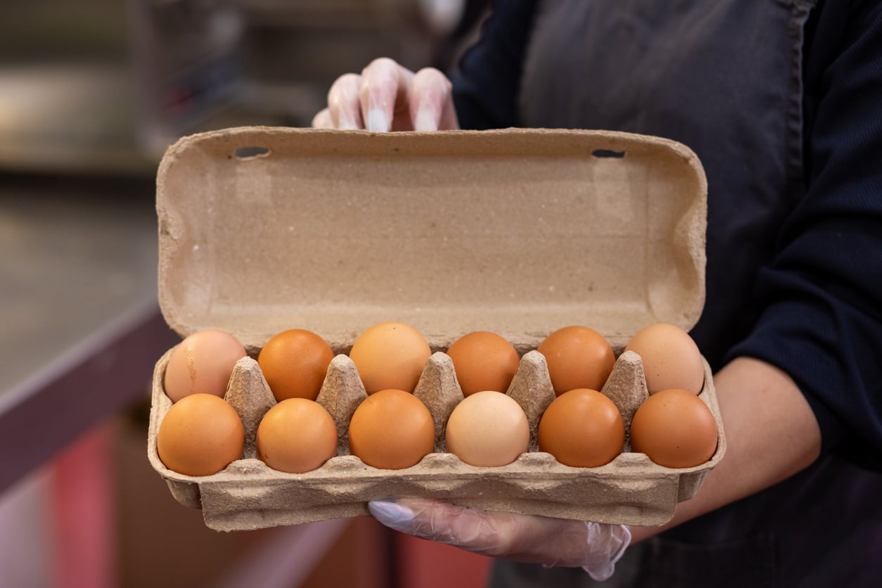 A person wearing a dark shirt and disposable glove holds an open carton of twelve brown eggs, with varying shades, in a cardboard egg container.
