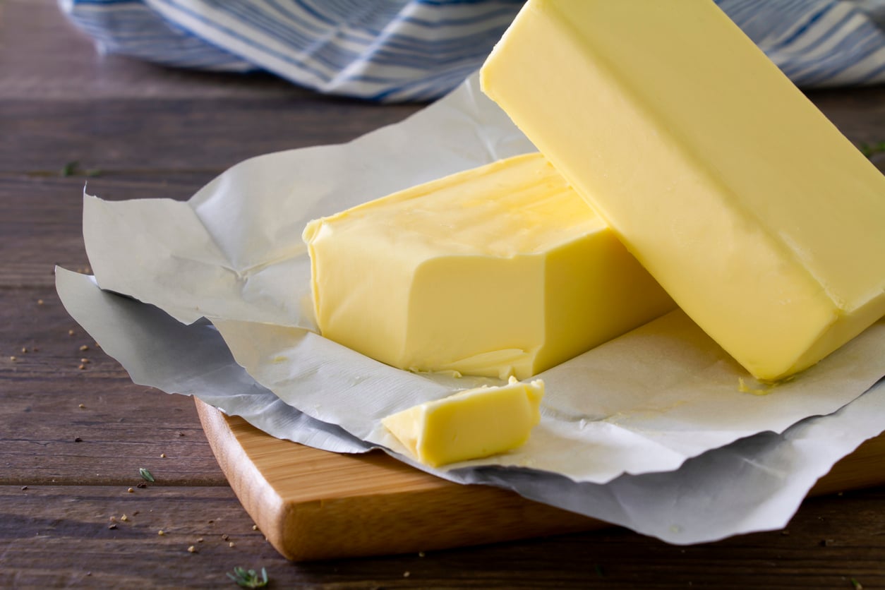 A block of butter partially unwrapped on parchment paper rests on a wooden cutting board, with a slice cut off and another block leaning against it. A blue and white striped cloth is in the background.