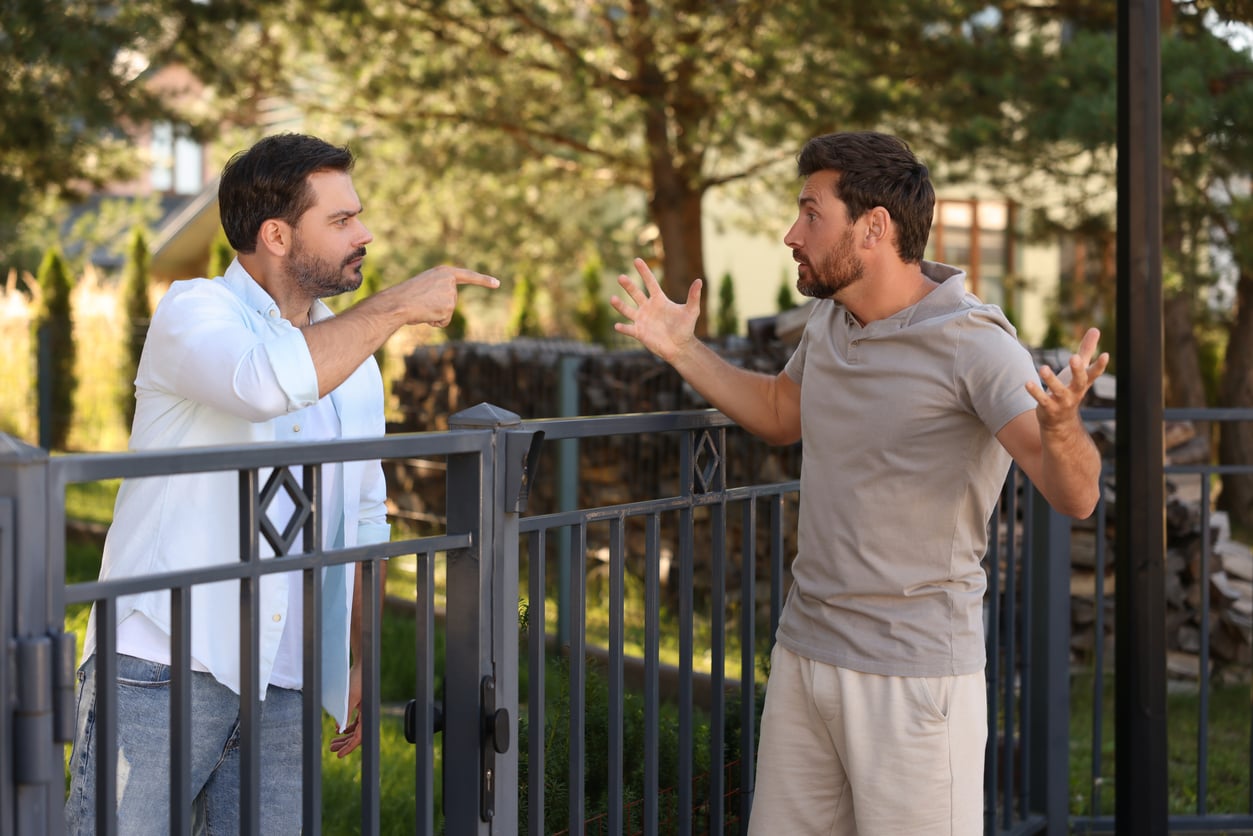 Two men standing on opposite sides of a metal fence are having an argument. One is pointing angrily, while the other gestures with his hands in frustration. Trees and greenery are visible in the background.