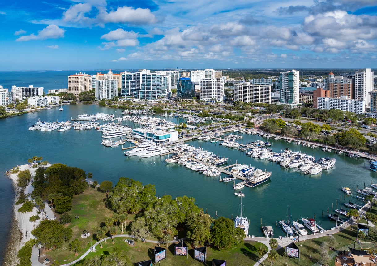 Aerial view of a marina filled with boats and yachts, surrounded by green parks and high-rise buildings under a blue sky with scattered clouds.