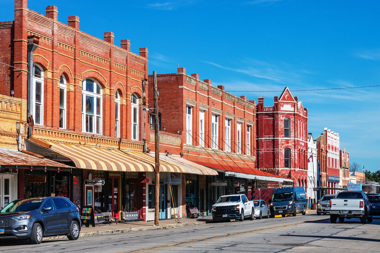 A row of historic red brick buildings with striped awnings lines a small town street, where cars and trucks are parked along the curb under a clear blue sky.