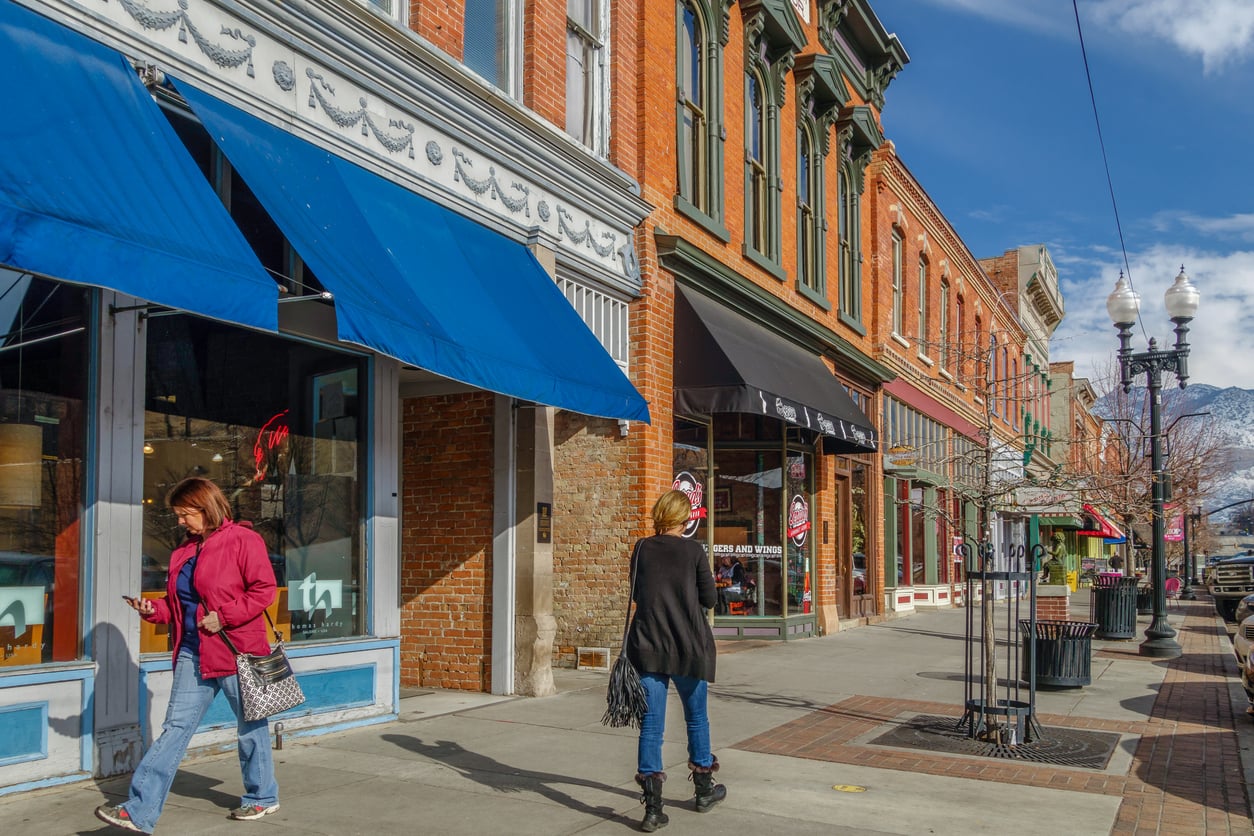 Two women stroll along a sunny downtown sidewalk in one of the Small Business States, surrounded by historic brick buildings, colorful awnings, shop windows, and leafless trees lining the vibrant street.