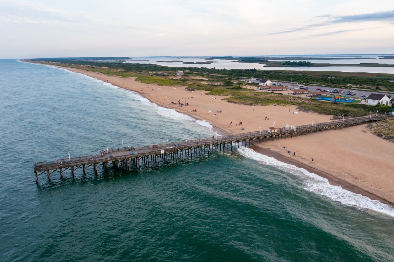 Aerial view of a wooden pier extending into the ocean, sandy beach with people walking, green vegetation, and several buildings on the shoreline under a partly cloudy sky.
