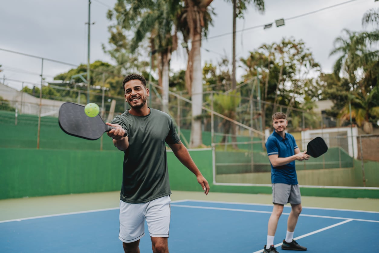 Two men enjoying one of the best budget-friendly hobbies, pickleball, on an outdoor court. The smiling man in front is about to hit the ball with his paddle, while his friend stands ready. Palm trees and a green fence provide a scenic background.