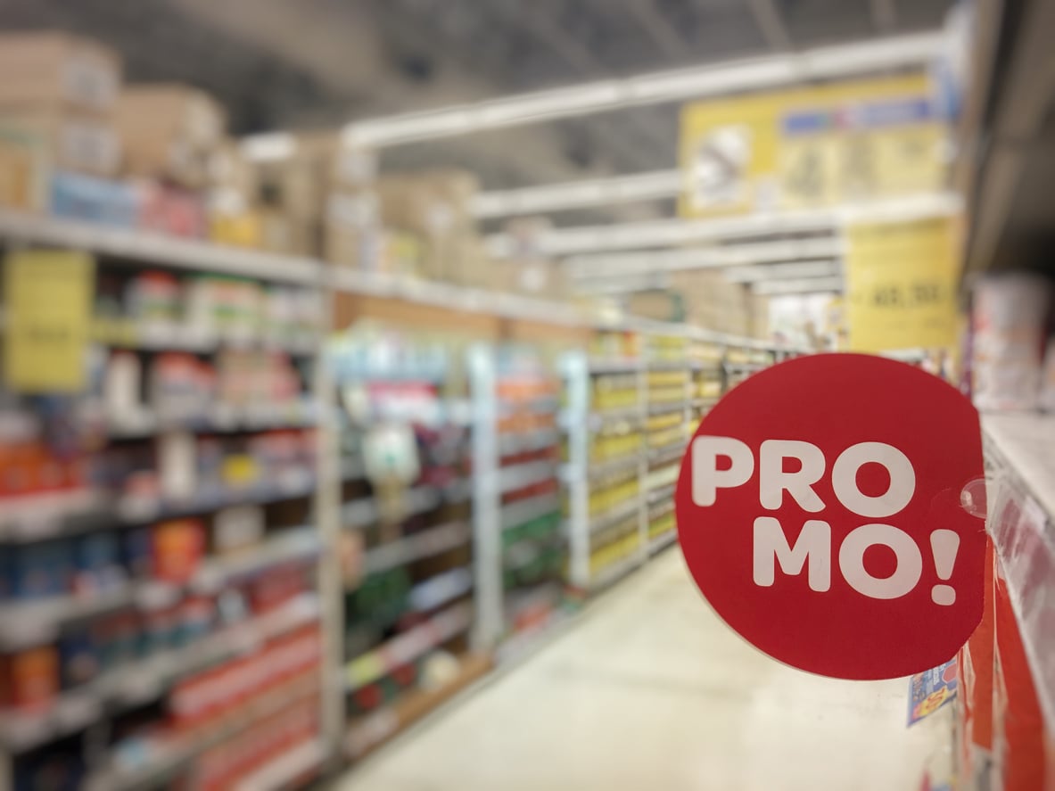 A red circular sign with white text reading "PROMO!" is attached to a supermarket shelf. The background shows blurred shelves filled with various products.