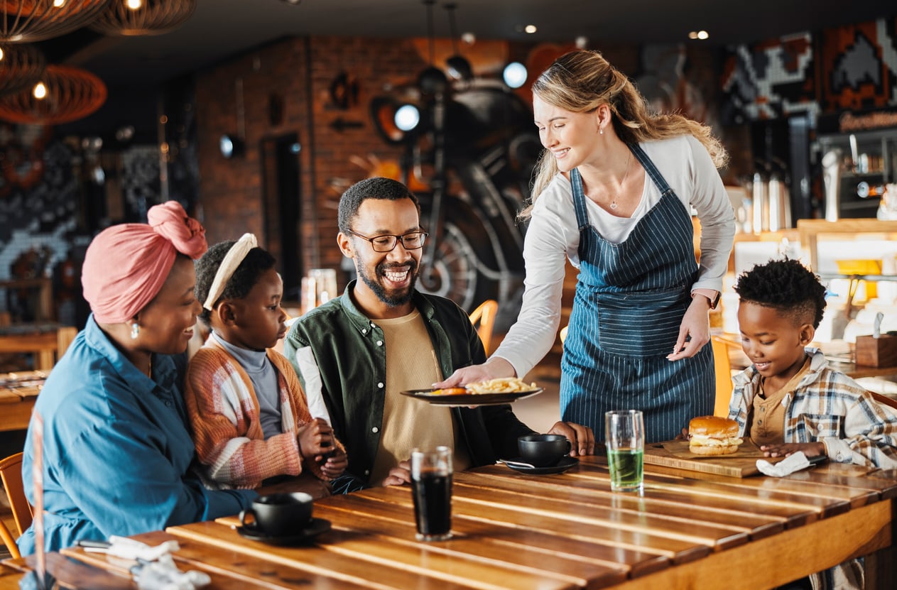 A smiling family of four sits at a wooden restaurant table as a waitress in an apron serves food. The parents and two children look happy and engaged, with food and drinks in front of them. The restaurant has a warm, modern decor.