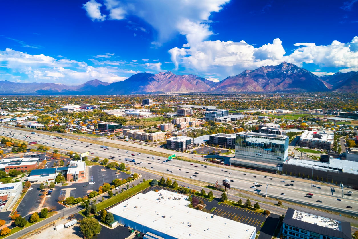 Aerial view of a city with a busy highway, modern buildings, and parking lots, set against a backdrop of mountains under a blue sky with scattered clouds.
