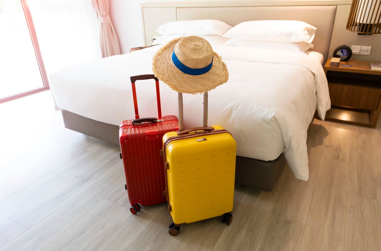 Two suitcases, one red and one yellow, stand upright on the floor in front of a neatly made bed. A straw hat with a blue ribbon rests on the handles. The room is bright with light wood floors.