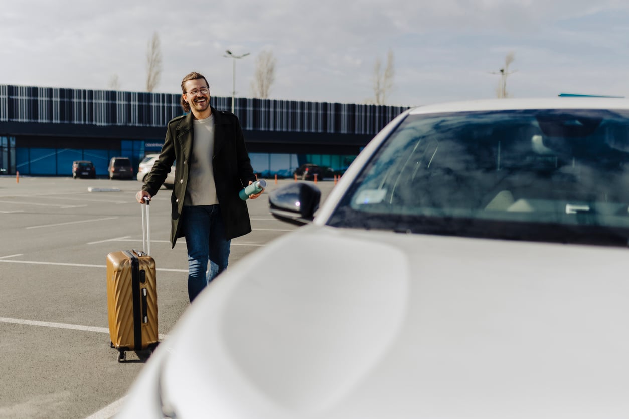 A smiling man with glasses walks through a parking lot, pulling a brown suitcase and holding a coffee cup, ready to share his Vegas money-saving tips as he approaches a white car on a sunny day.