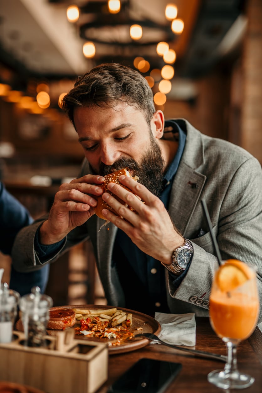 A man with a beard, wearing a gray blazer, sits at a restaurant table enjoying a big bite of a hamburger—maybe savoring one of those products men skip buying at home. Fries, salad, and an orange drink sit nearby. The background is warmly lit and out of focus.