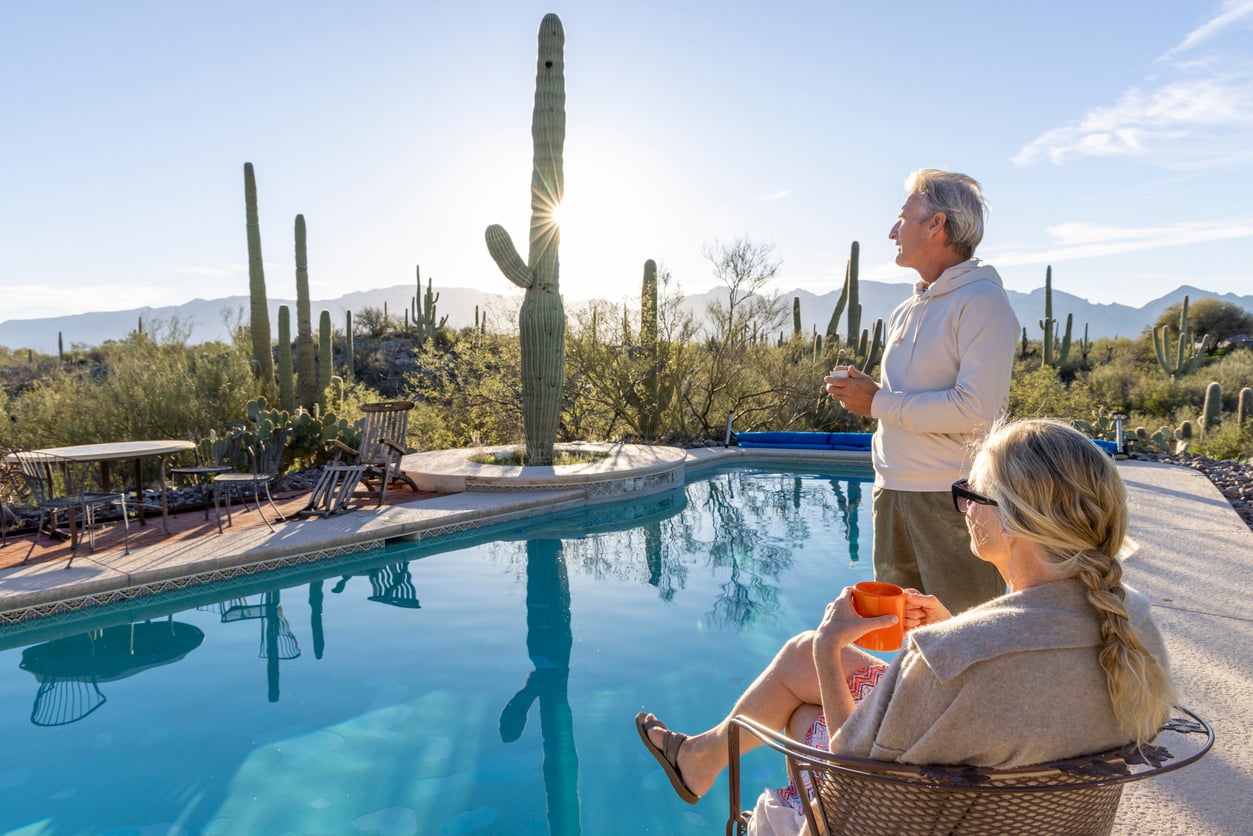 An older man and woman relax by a pool in a desert setting, surrounded by cacti, as the sun rises or sets over distant mountains. The woman sits with a mug, while the man stands looking at the view.