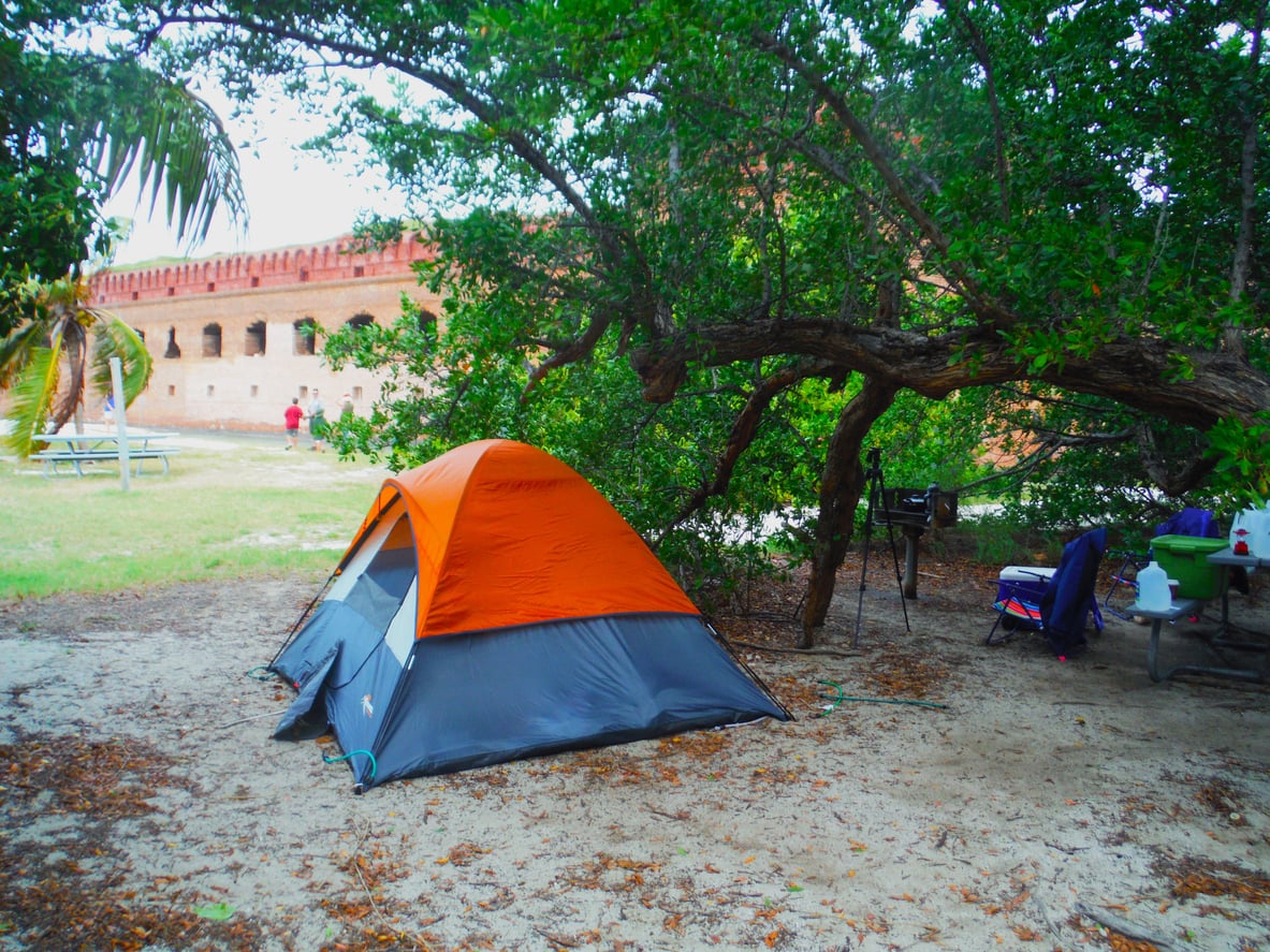 A small orange and gray tent is set up under a leafy tree near a brick fort. There’s a picnic table, grill, and camping gear in the background on sandy ground.