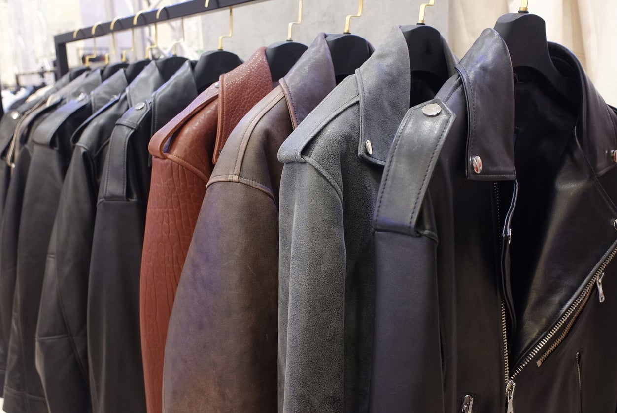 A row of leather jackets in various colors, including black and brown, hanging neatly on black hangers among other stylish clothes in a clothing store.