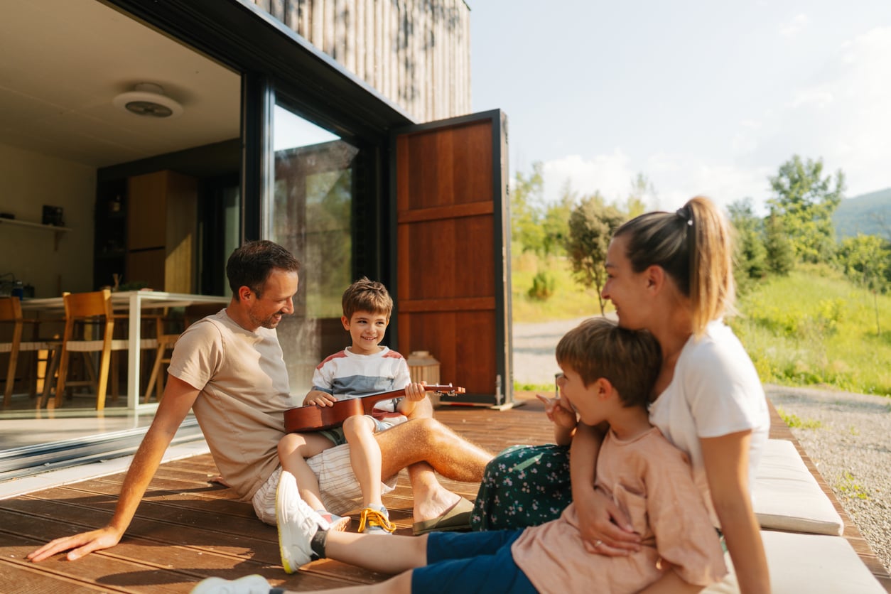A family of four sits on a wooden deck outside their home on a sunny day. The father and son smile at each other while the son holds a ukulele. The mother and younger child sit close together, all looking happy and relaxed.